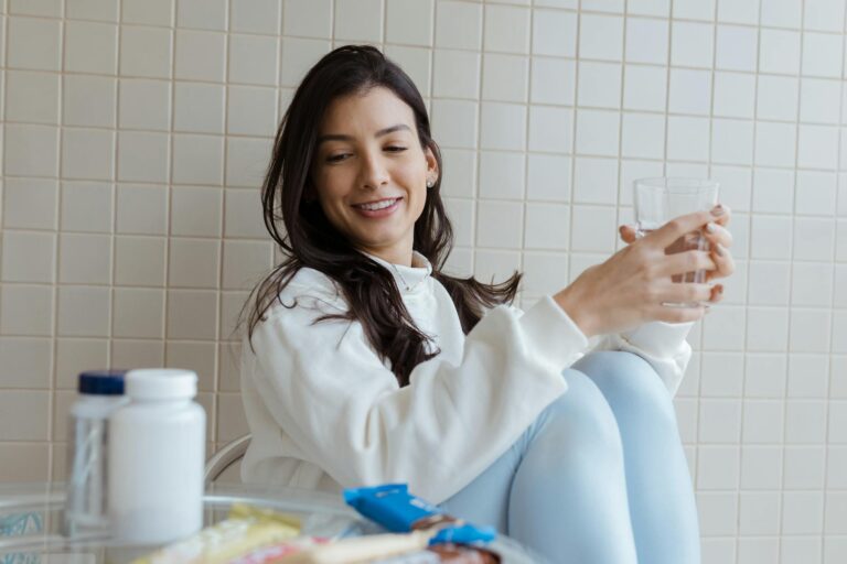 Smiling woman holding a glass while seated, with pill bottles on the table.