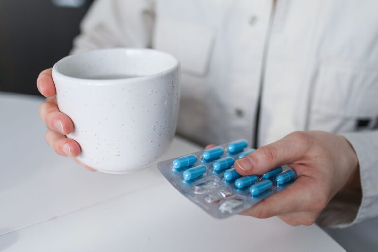 Close-up of a woman holding a hot drink and blister pack of blue capsules indoors, suggesting illness or treatment.