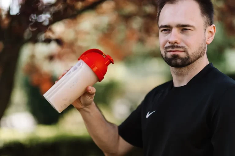 Adult man holds a protein shake in a red lid tumbler outdoors, wearing a black shirt.